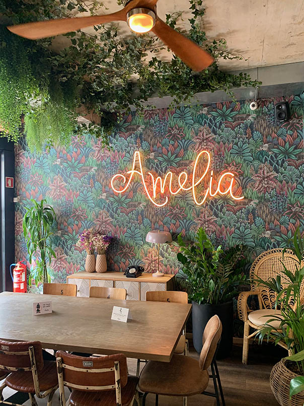 interior de la cafetería Amelia brunch mobiliario vintage pared florecida logo rosa neón verde y plantas colgantes