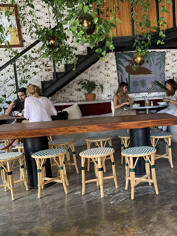 interior of the Janis café in the centre large solid wood table rattan stools in the background two couples having lunch on a sofa metal stairs hanging plants 