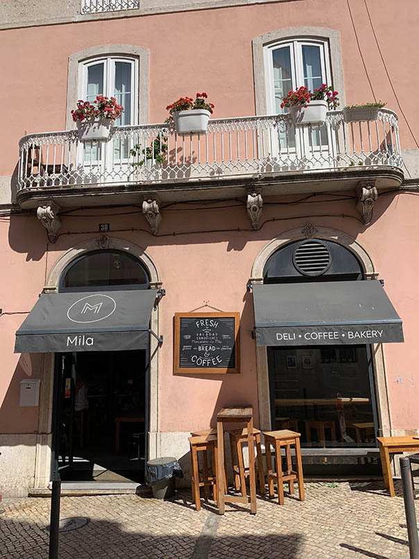 Mila winkelpui en café roze gevel met wit balkon zwarte luifel met logo erop en een lei met vers brood en koffie in wit krijt