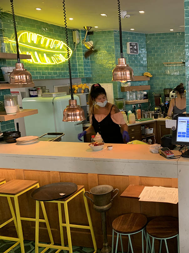 inside the Nicolau café two employees preparing orders behind a counter the walls are in turquoise blue earthenware copper and neon lamps in the shape of yellow feather