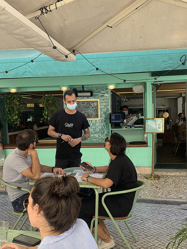 terras met klanten die een praatje maken met de ober tafels en stoelen en de voorgevel van het café-restaurant in groen-blauwe kleur    